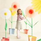 Young girl posing in a studio setting surrounded by large, colorful stand-up paper flower decorations.
