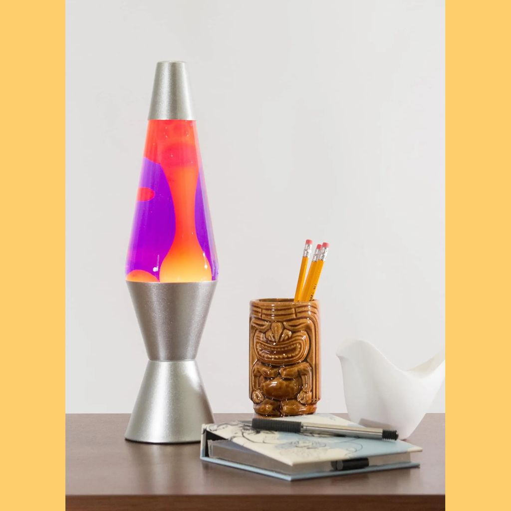 Silver lava lamp with purple and orange wax sits on a wooden table. Next to it, a tiki mug holds pencils beside a notepad and pen, with a serene white dove figure nearby.