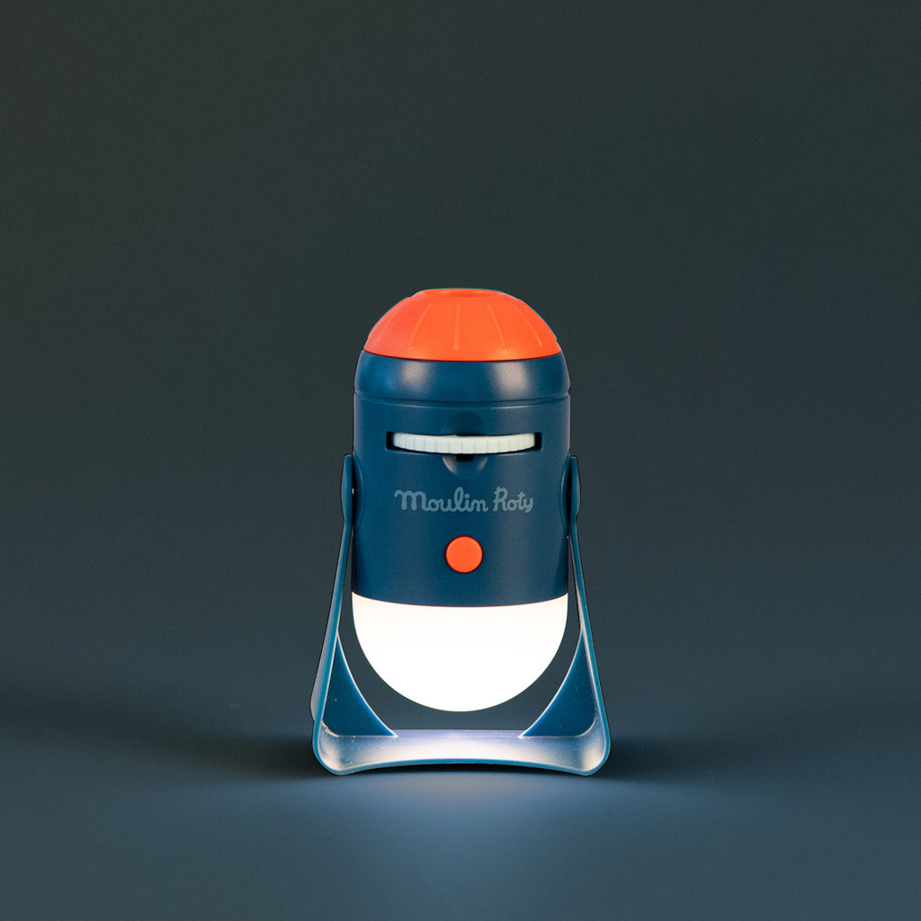 A blue and orange toy projector titled "Moulin Roty" emits a focused beam of light. It stands on a gray surface, giving a playful and whimsical tone.