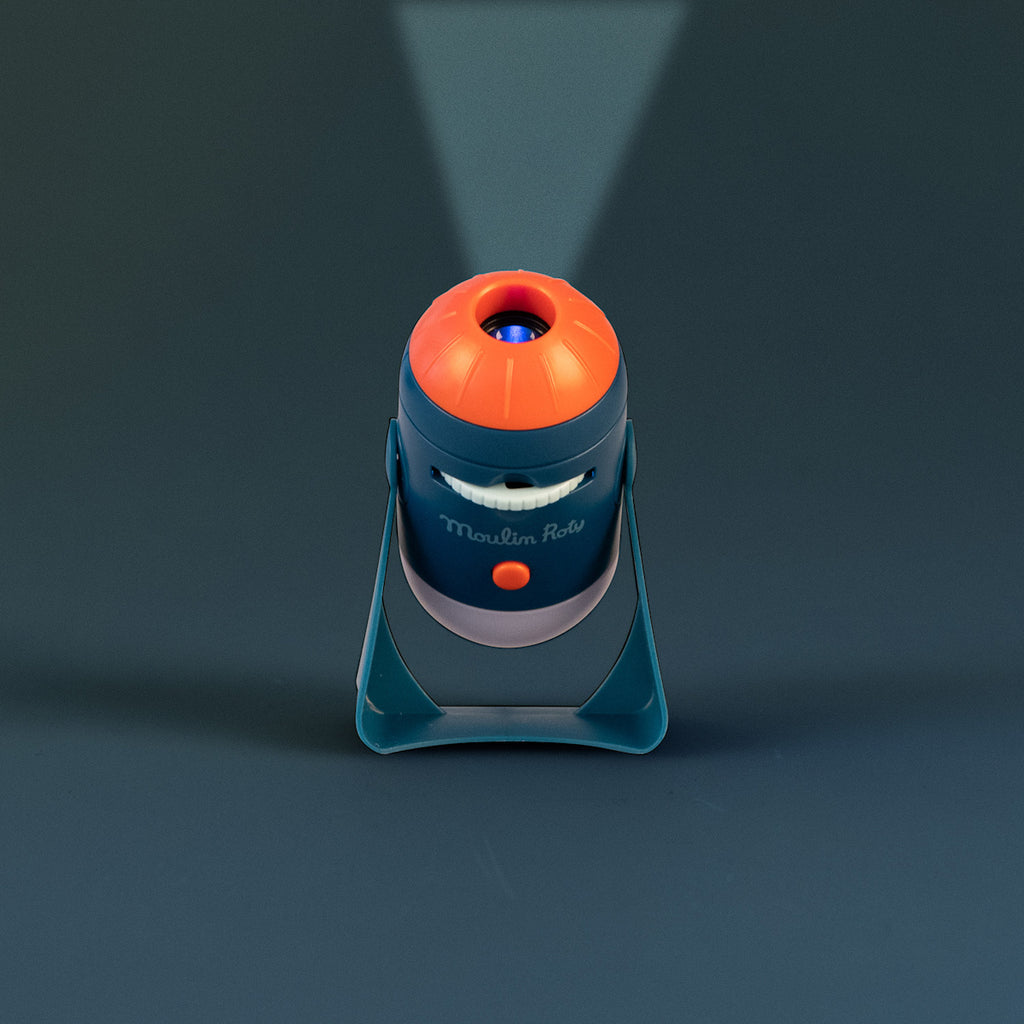 A blue and orange toy projector titled "Moulin Roty" emits a focused beam of light. It stands on a gray surface, giving a playful and whimsical tone.