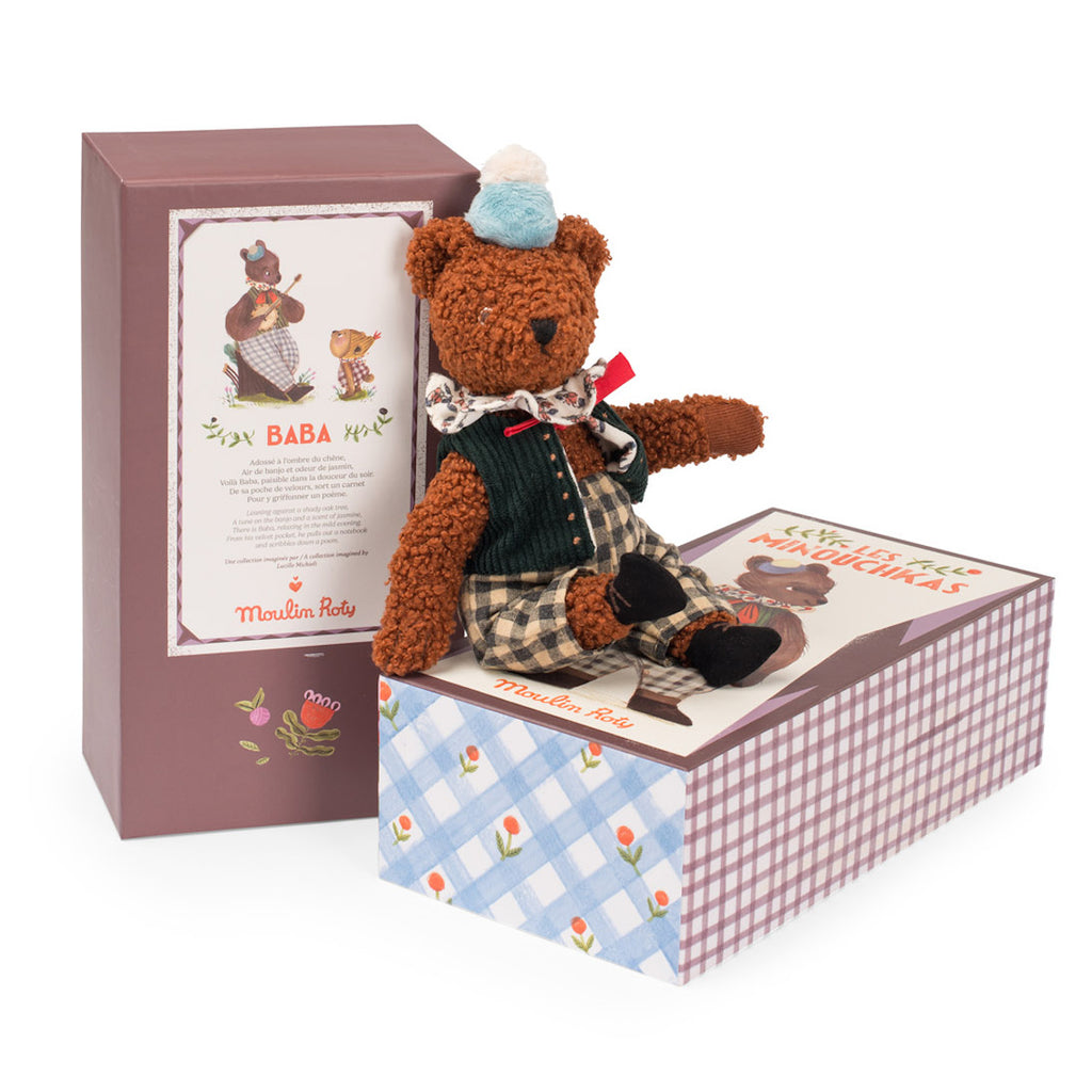 A plush teddy bear in a green vest, checkered pants, and a blue hat sits on a decorative box. Another box is beside it, both colorful and patterned.