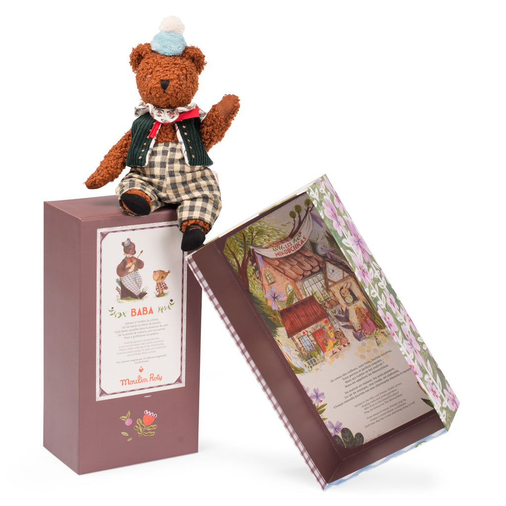 A plush teddy bear in a green vest, checkered pants, and a blue hat sits on a decorative box. Another box is beside it, both colorful and patterned.