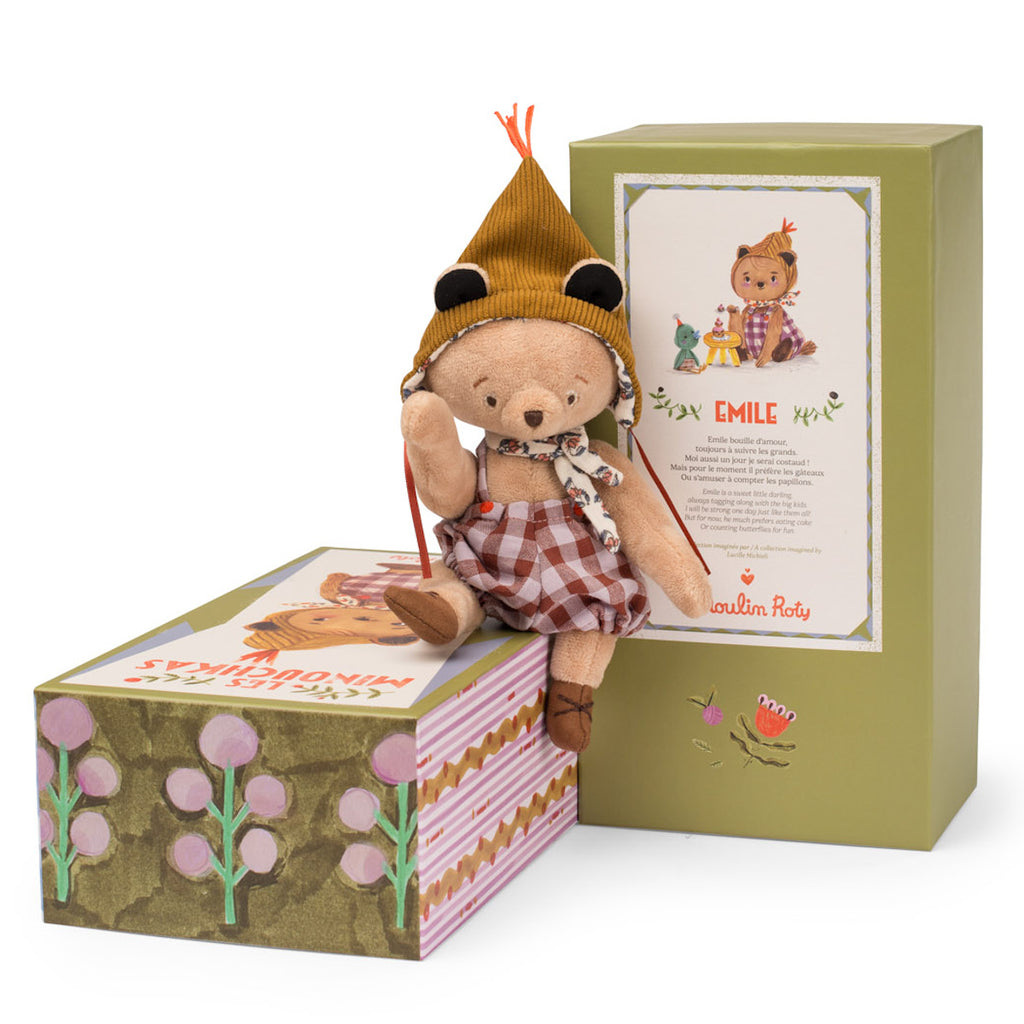 A plush bear wearing a brown checked dress and yellow knit hat sits on colorful, illustrated packaging. The mood is playful and whimsical.