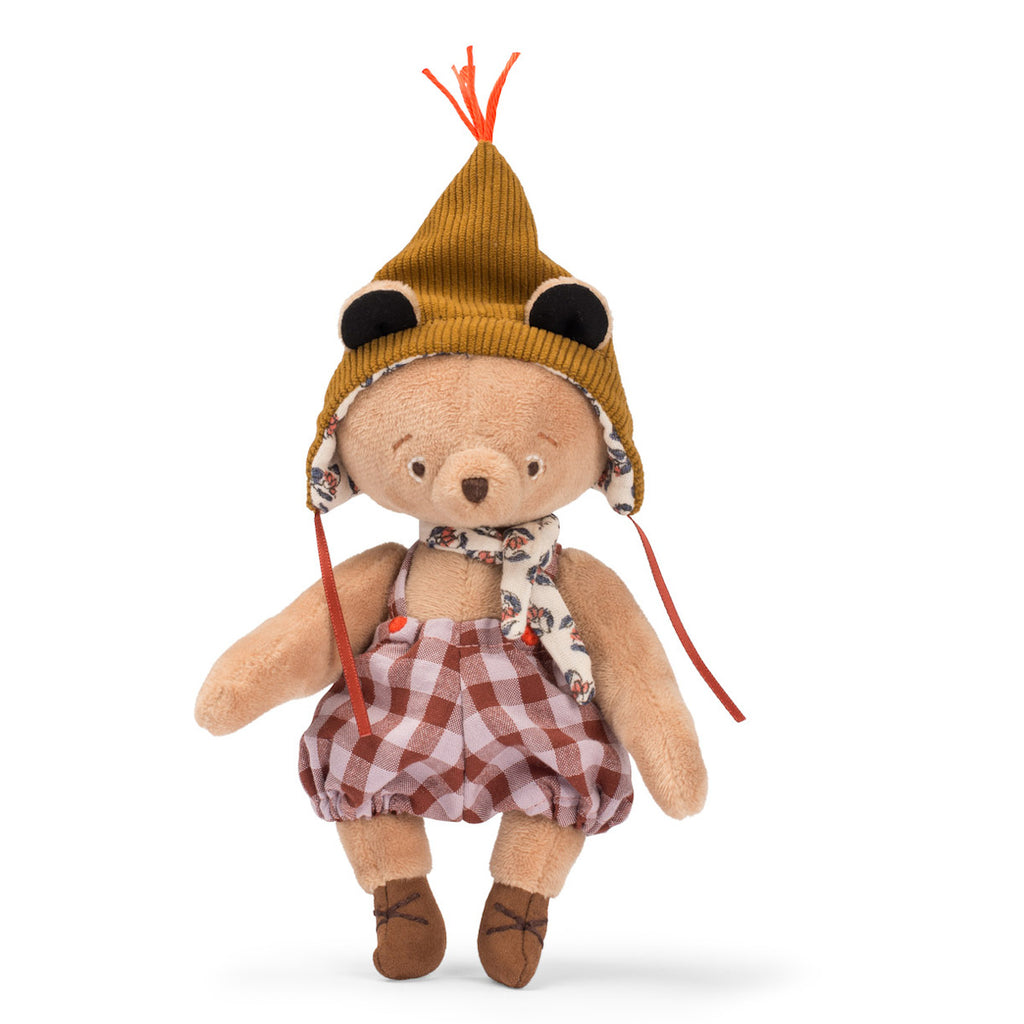 Stuffed bear in checkered dress, floral scarf, and brown hat with orange tassel and earflaps. Its cozy, whimsical attire conveys playful charm.