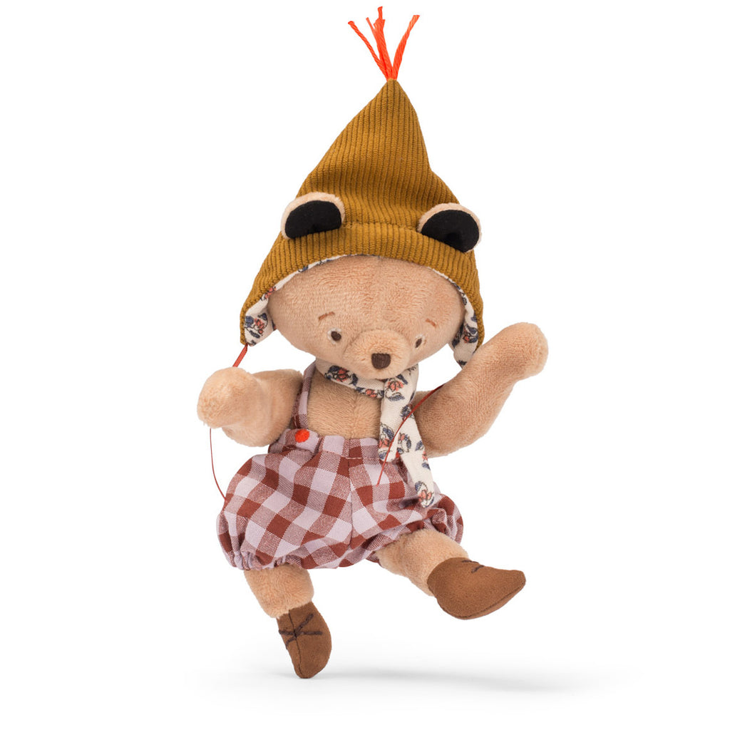 Stuffed bear in checkered dress, floral scarf, and brown hat with orange tassel and earflaps. Its cozy, whimsical attire conveys playful charm.