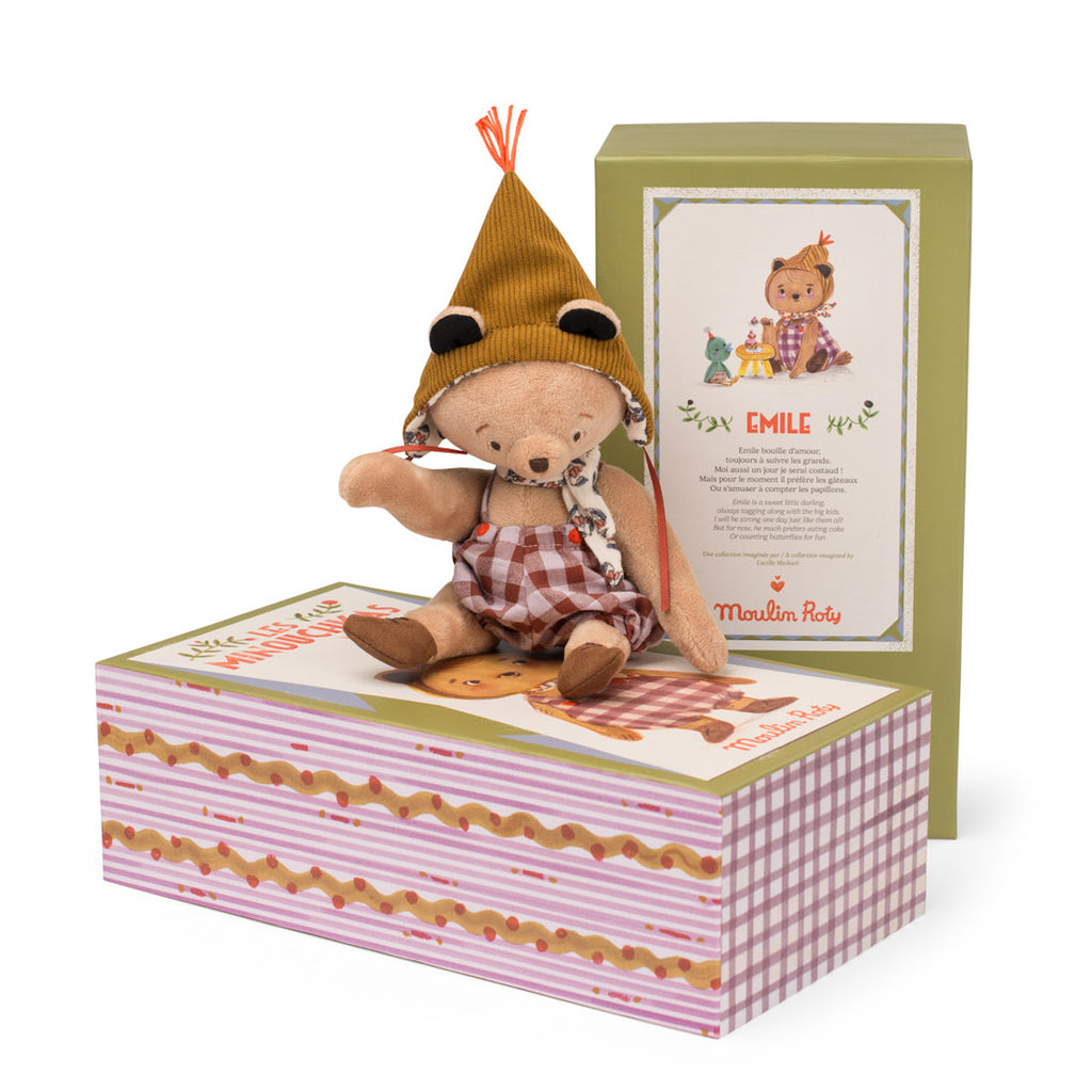 A plush bear wearing a brown checked dress and yellow knit hat sits on colorful, illustrated packaging. The mood is playful and whimsical.