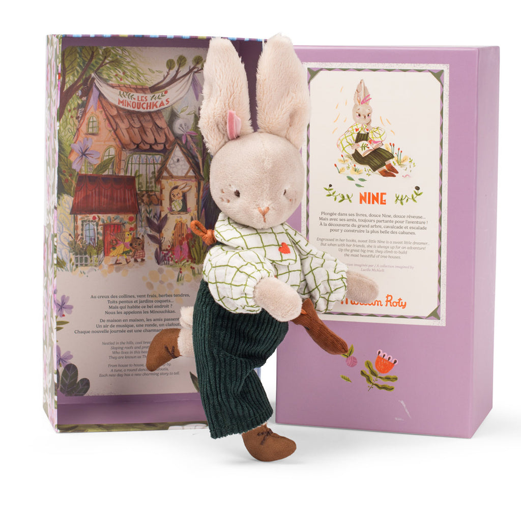 A plush rabbit toy wearing a checkered shirt, green corduroy pants, and a small brown satchel sits by a decorative box. The box features floral designs and text. The scene is playful and charming.