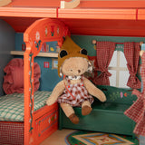 Plush toy bear in a pointed hat sitting inside a decorated wooden toy caravan next to a mini bed.