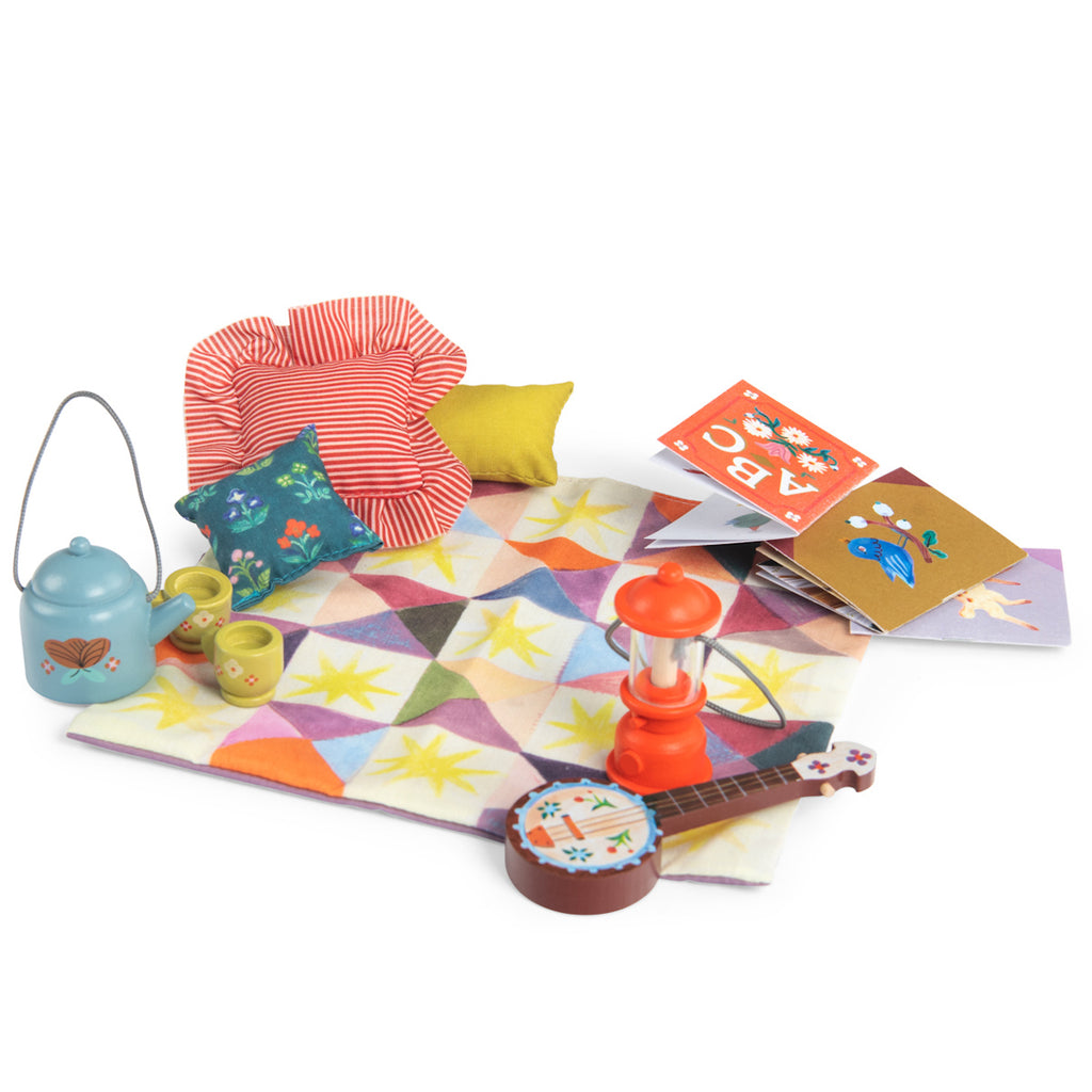 Cozy picnic scene with a colorful quilt, red lantern, teal teapot, cups, and vibrant pillows. Nearby are illustrated books and a small banjo.