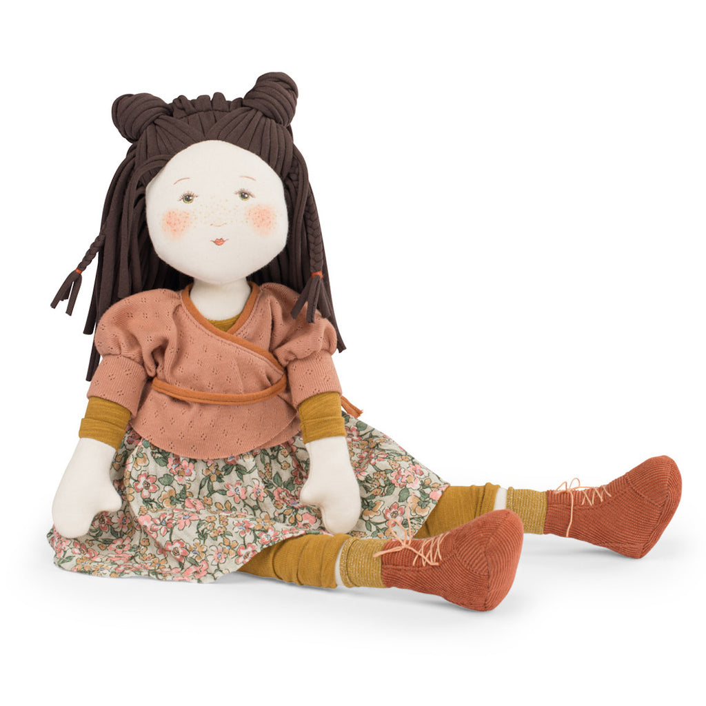 A plush doll with brown braided hair in two buns, wearing a peach top, floral skirt, yellow socks, and orange shoes. The doll's face is cheerful.