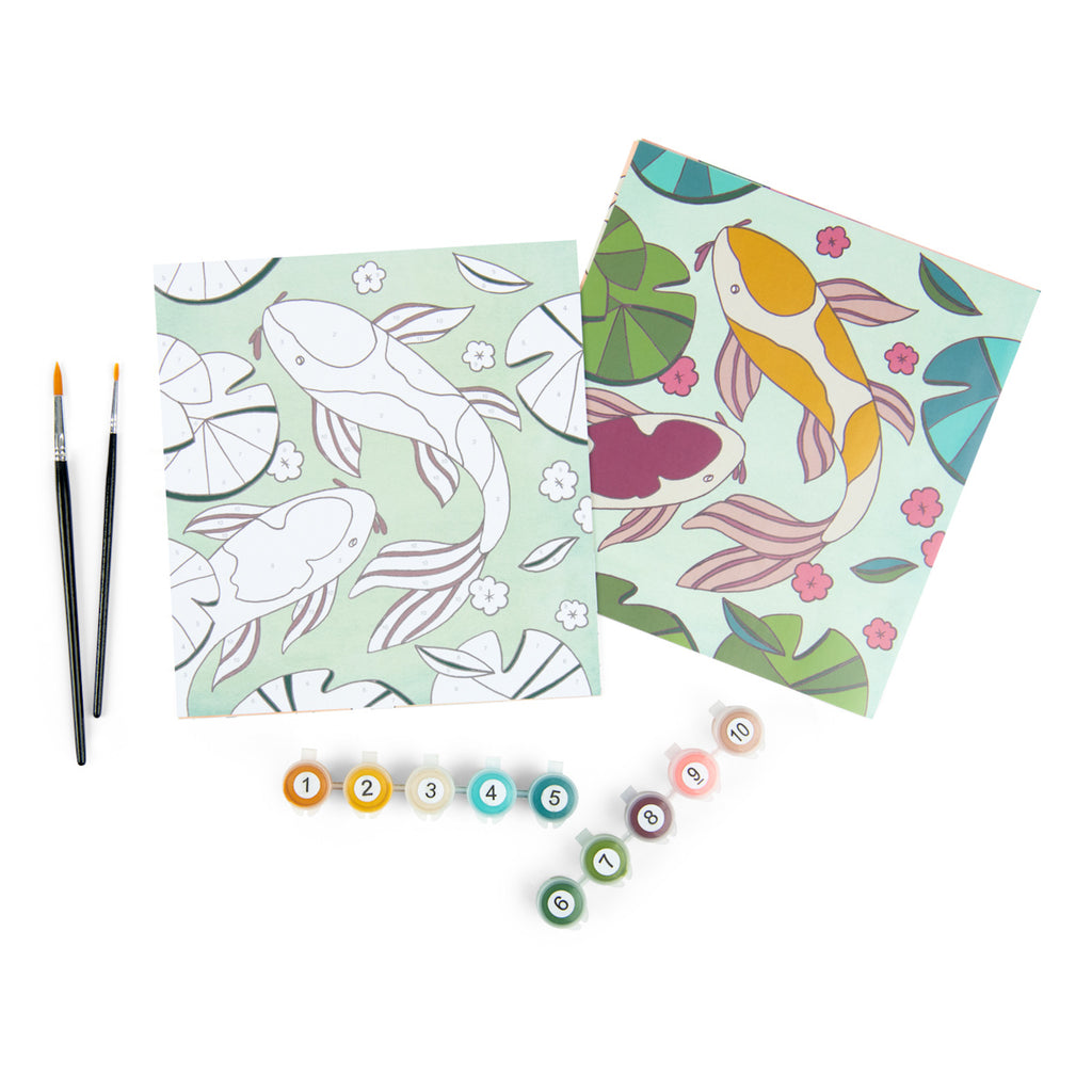 Two paint-by-number canvases depict koi fish and lily pads against a light green background. Nearby are paintbrushes and numbered paint pots.