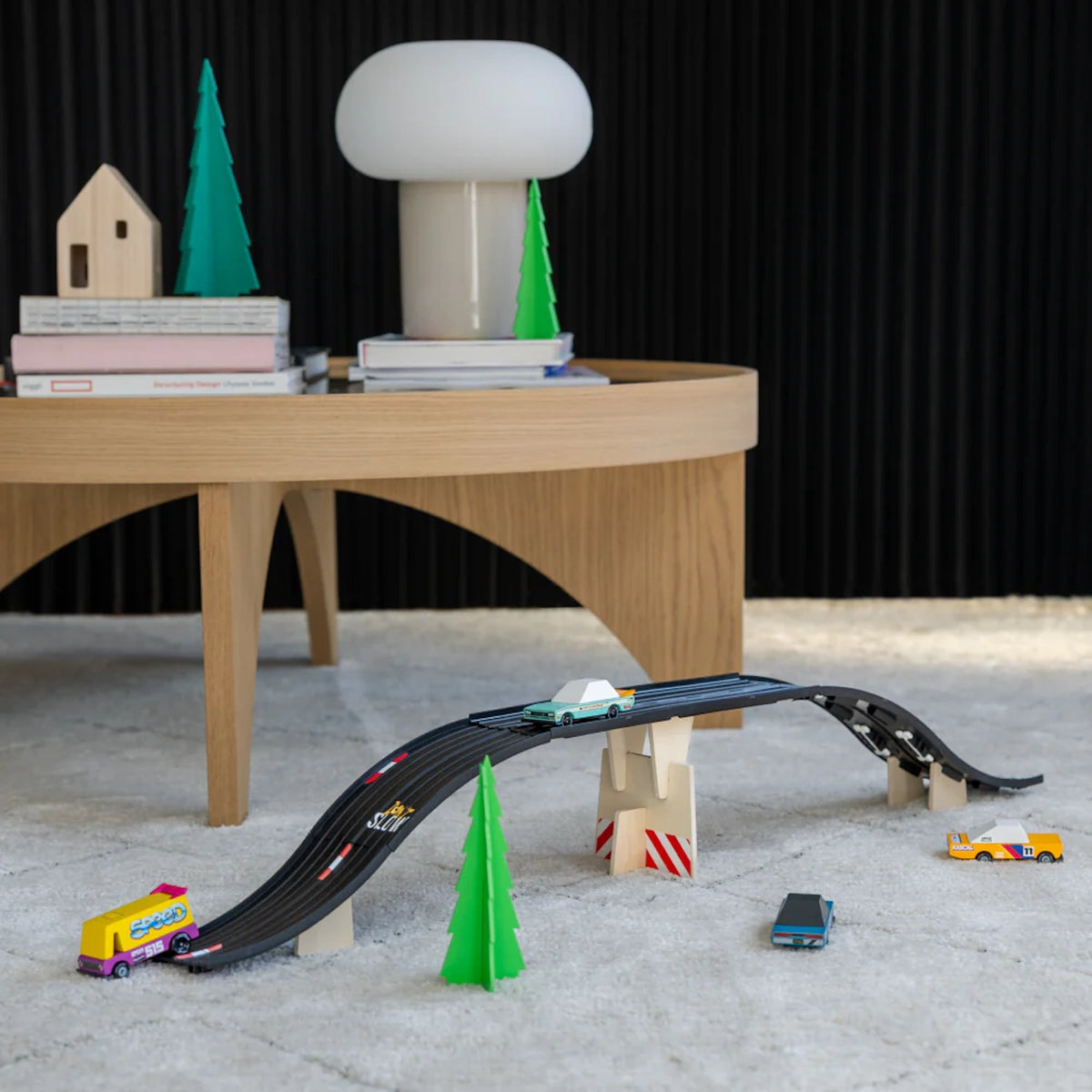 A toy car playset with a black track bridge, miniature cars, trees, and a modern table in the background.