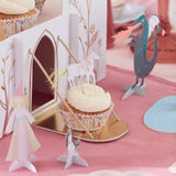 A cupcake with a unicorn topper sits on a toy castle drawbridge, surrounded by paper dragons and princesses.