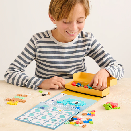 A child builds a blue ocean creature with Plus-Plus blocks using a spiral-bound activity pad and a yellow storage box.