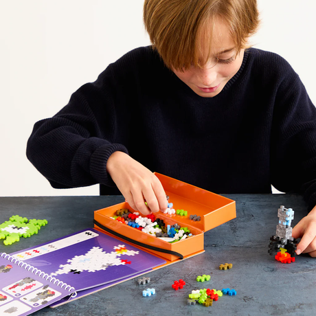 Child building a space-themed Plus-Plus puzzle using a toy activity pad, orange storage box, and colorful 2D and 3D pieces.