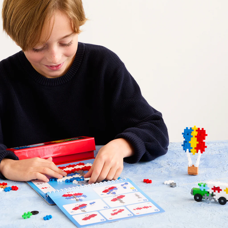 Child building a transportation Plus-Plus puzzle with a toy activity pad, red storage box, and colorful 2D and 3D pieces.