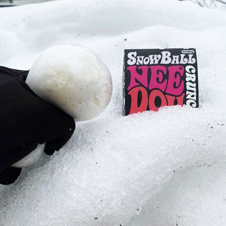 A person in a black glove squeezes a Schylling Snow Ball Crunch Nee-Doh stress ball in real snow next to its packaging.