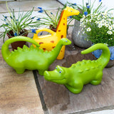 Yellow giraffe and green dinosaur animal-shaped watering cans for kids on a stone patio next to potted garden plants.