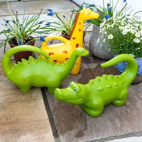 Yellow giraffe and green dinosaur animal-shaped watering cans for kids on a stone patio next to potted garden plants.