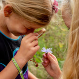 Child using a wooden magnifying glass to examine a purple wildflower held by a friend outdoors.