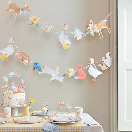 A birthday party table setting featuring woodland animal garlands, a floral cake, and matching tableware.