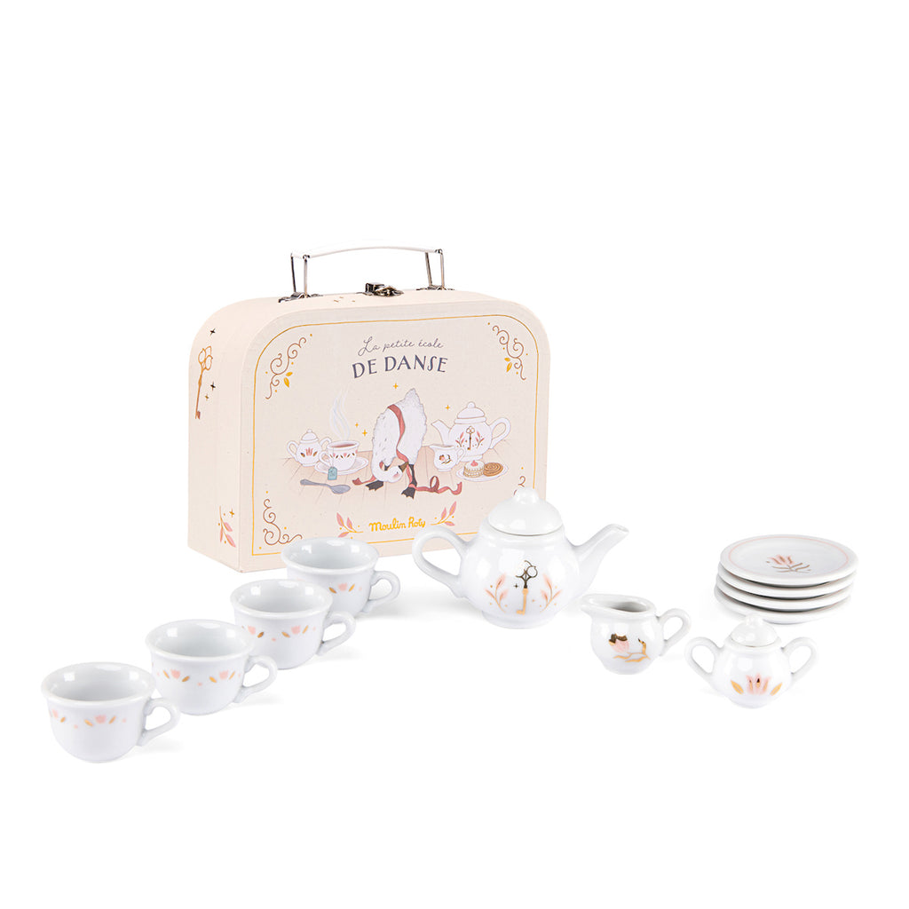 A children's tea set with white ceramic cups, teapot, and plates featuring delicate designs. A suitcase with "Le petite table De Danse" is in the background.