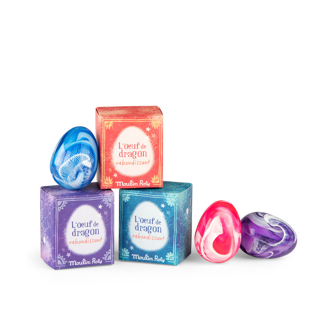 Three colorful boxes with "L'oeuf de dragon rebondissant" text are stacked with three marble-patterned eggs in blue, pink, and purple beside them. Playful and magical tone.