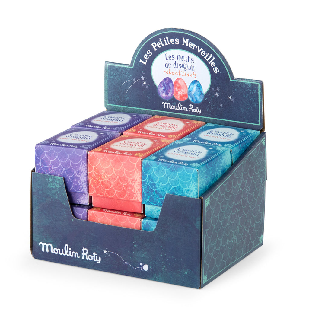 Display box of colorful "Moulin Roty Les Petites Merveilles" dragon eggs. Packaging features purple, red, and blue scaled designs, conveying a magical theme.