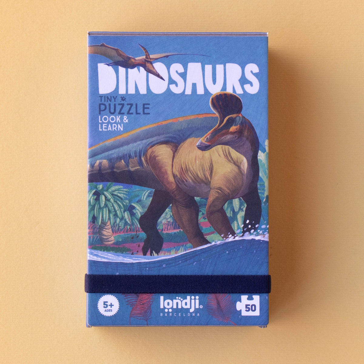 Box of a dinosaur-themed puzzle for ages 5+. Features a dinosaur and flying reptile illustration with tropical plants, set against a vibrant blue background.