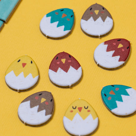 Colorful wooden Piu Piu counting game pieces shaped like chicks hatching from white eggshells on a yellow background.