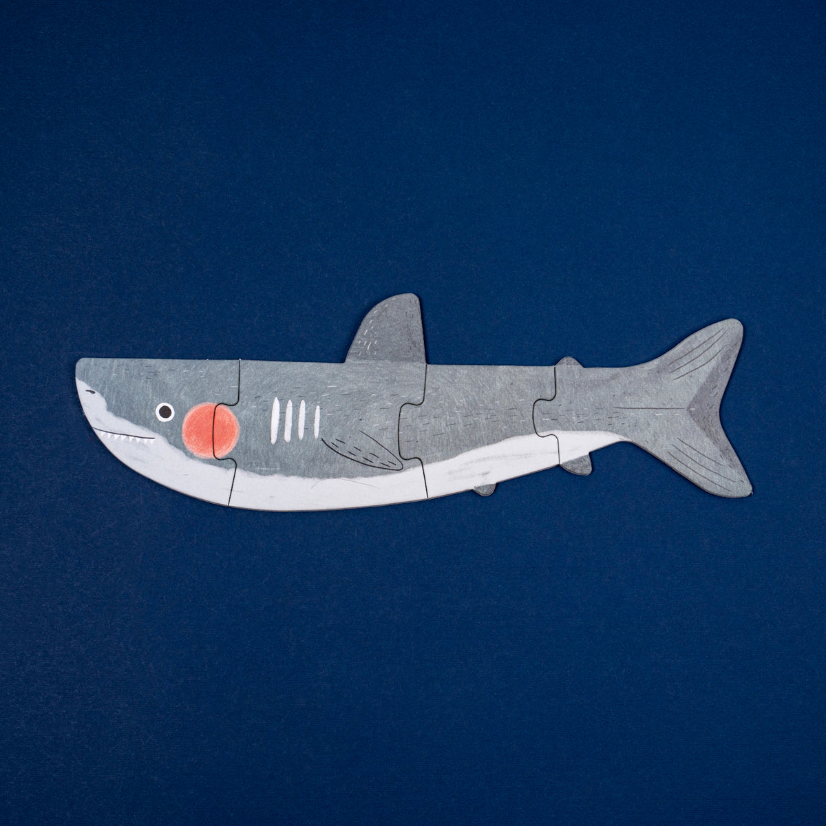 A Londji wooden shark puzzle featuring a grey shark with a red cheek, composed of four pieces on a dark blue background.