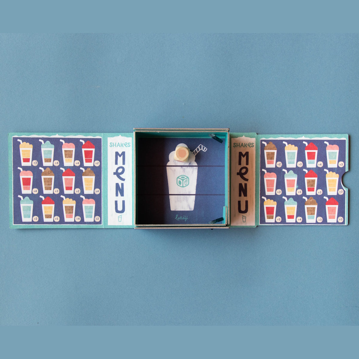 Londji Shakes tiny board game with colorful milkshake menu cards, a wooden die, and a blue illustrated box.