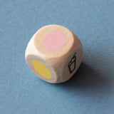 Wooden die with pink and yellow circles and a milkshake icon on a blue background. Londji tiny game shakes wooden dice.
