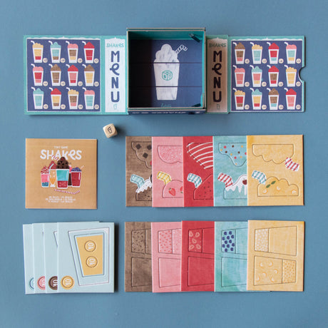 Londji Shakes tiny board game with colorful milkshake cards, menus, dice, and box on a blue background.