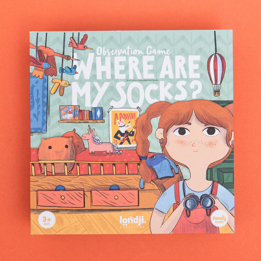 Illustrated game box cover with a girl holding binoculars, toys, and text "Where Are My Socks?" on a patterned background. Whimsical and playful.