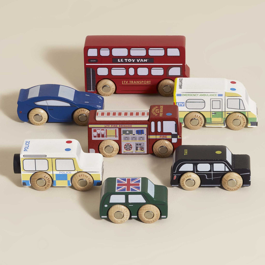 Toy wooden vehicles including a red double-decker bus, police car, ambulance, fire truck, green car, and black taxi. Playful and colorful.