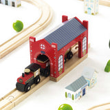 Wooden toy train exiting a red locomotive shed on tracks with miniature houses nearby.