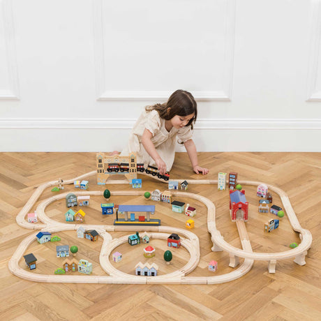 Young girl playing with a large wooden train set on a wood floor in front of a white wall.