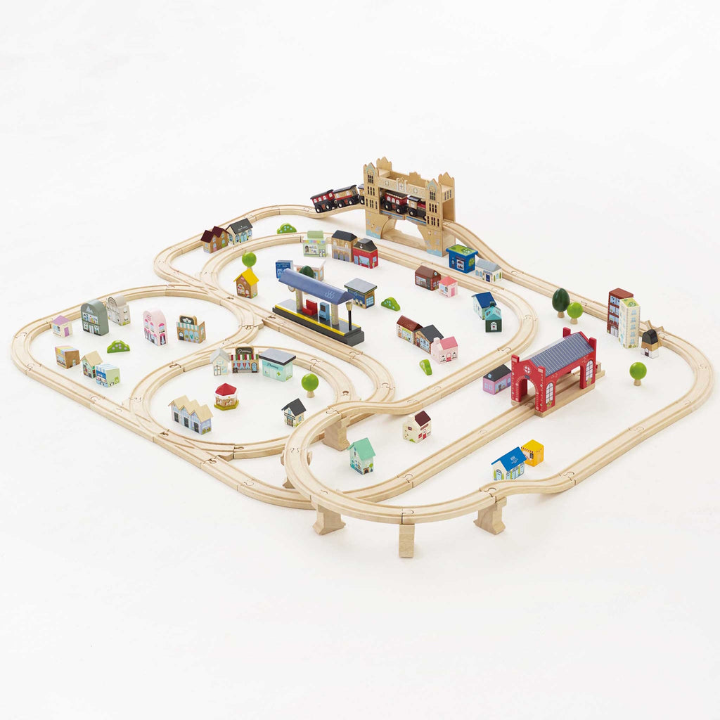 Wooden train set on white background with tracks forming loops and curves. Colorful miniature buildings and trees are scattered around, creating a playful town scene.