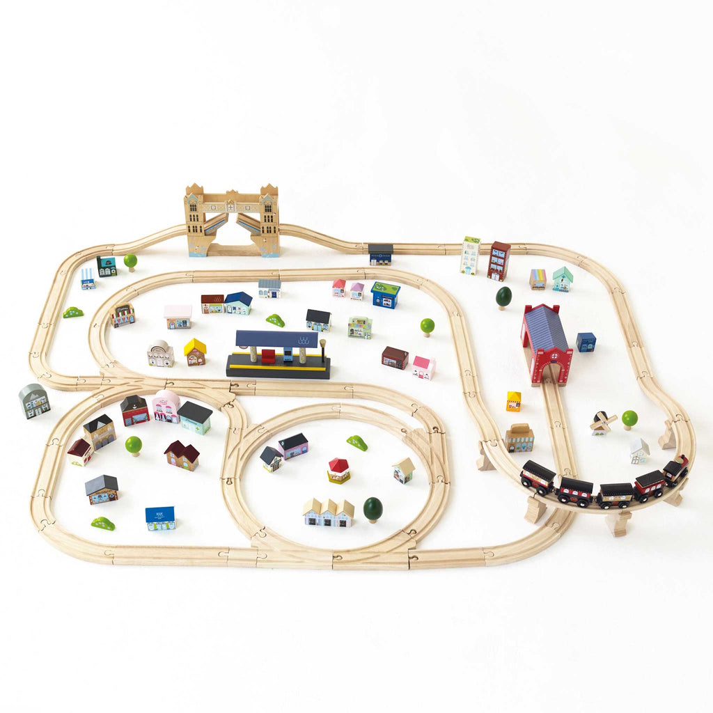 Wooden train set on white background with tracks forming loops and curves. Colorful miniature buildings and trees are scattered around, creating a playful town scene.