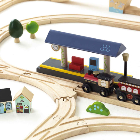 Wooden toy train stopped at a miniature station platform with tracks and small buildings.