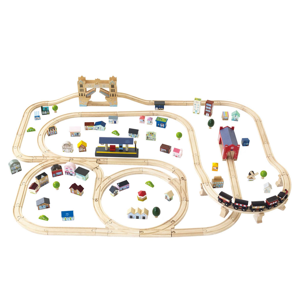 Wooden train set on white background with tracks forming loops and curves. Colorful miniature buildings and trees are scattered around, creating a playful town scene.