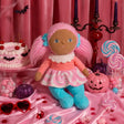 Plush doll with pink pigtails surrounded by pink Halloween decor, lollipops, and a cake.