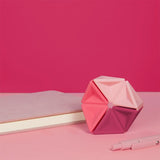 Pink silicone hexaflexagon fidget toy upright on a desk next to a book and pen.