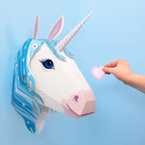 A hand holds a pink felt heart towards a paper unicorn head with blue mane and striped horn, mounted on a light blue wall, creating a whimsical feel.