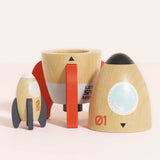 Small wooden rocket standing next to the base and nose cone of a larger toy space rocket set.