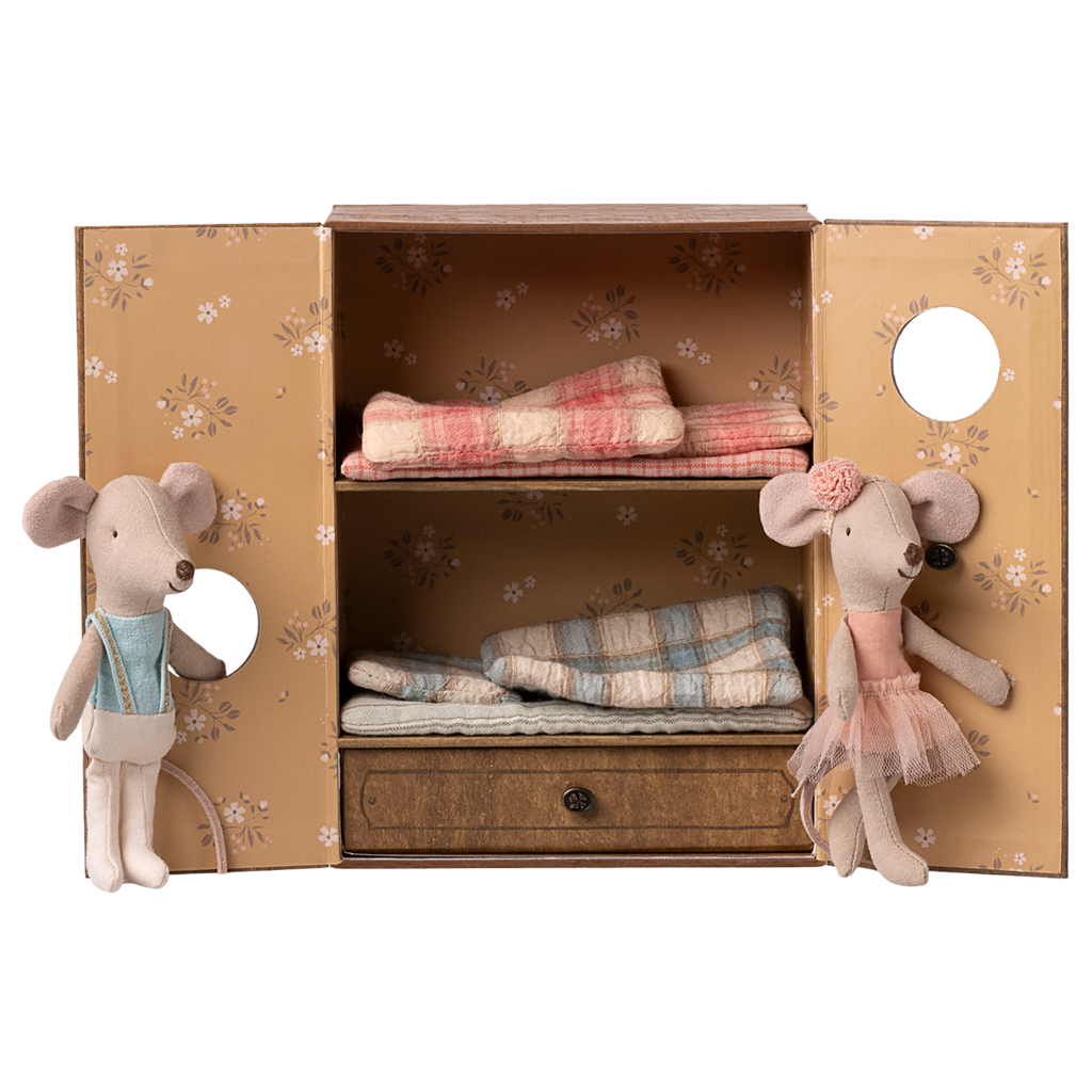 Fabric mice figures dressed in overalls and a tutu stand beside a floral-patterned cabinet. Inside are folded plaid blankets, creating a cozy, playful scene.