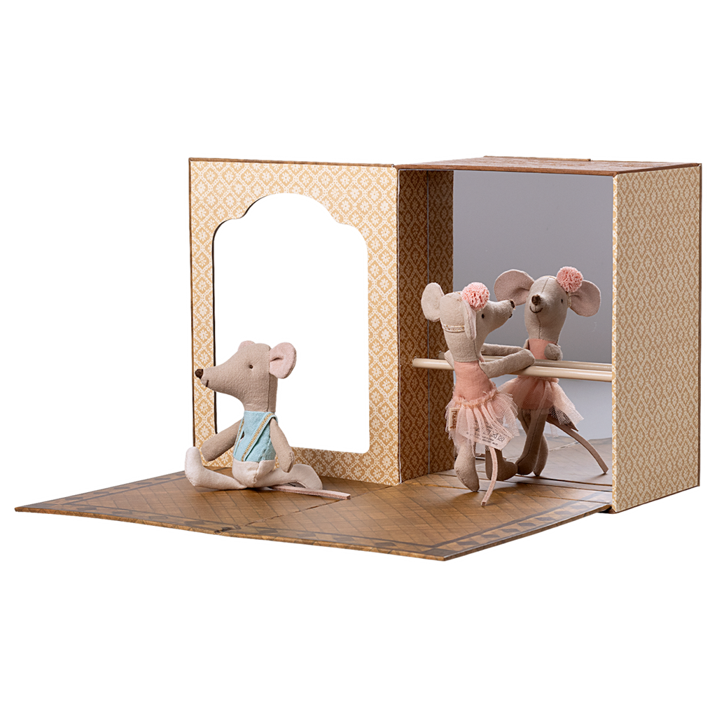 Three plush mice are in a ballet studio. Two mice in pink tutus pose at a barre, while a third sits nearby in blue attire, conveying a playful tone.