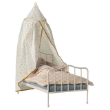 A miniature white metal bed with a blue-striped mattress and floral-patterned bedding. A white canopy with golden spots drapes over, creating a cozy, whimsical feel.