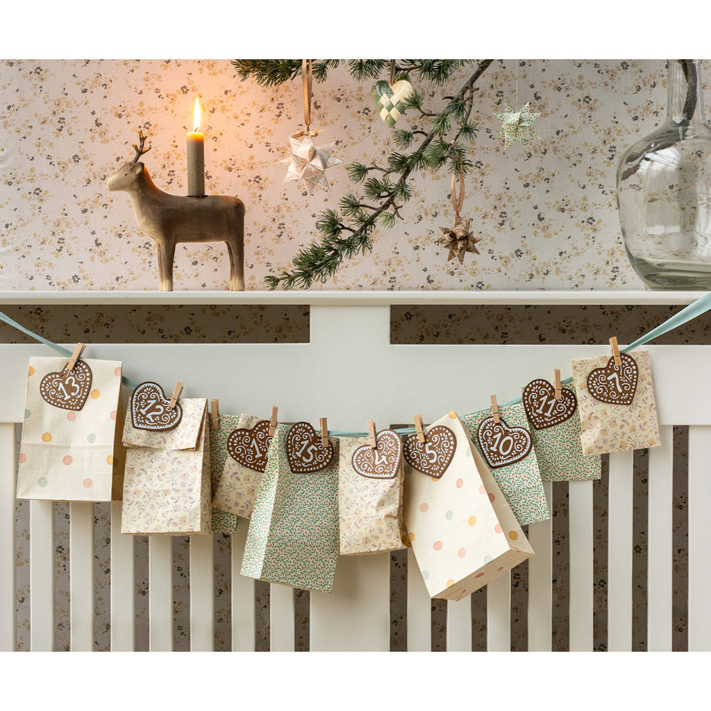Festive scene featuring a handmade Advent calendar with numbered heart-shaped cookies on a string. A reindeer candle and pine branch enhance the cozy holiday atmosphere.